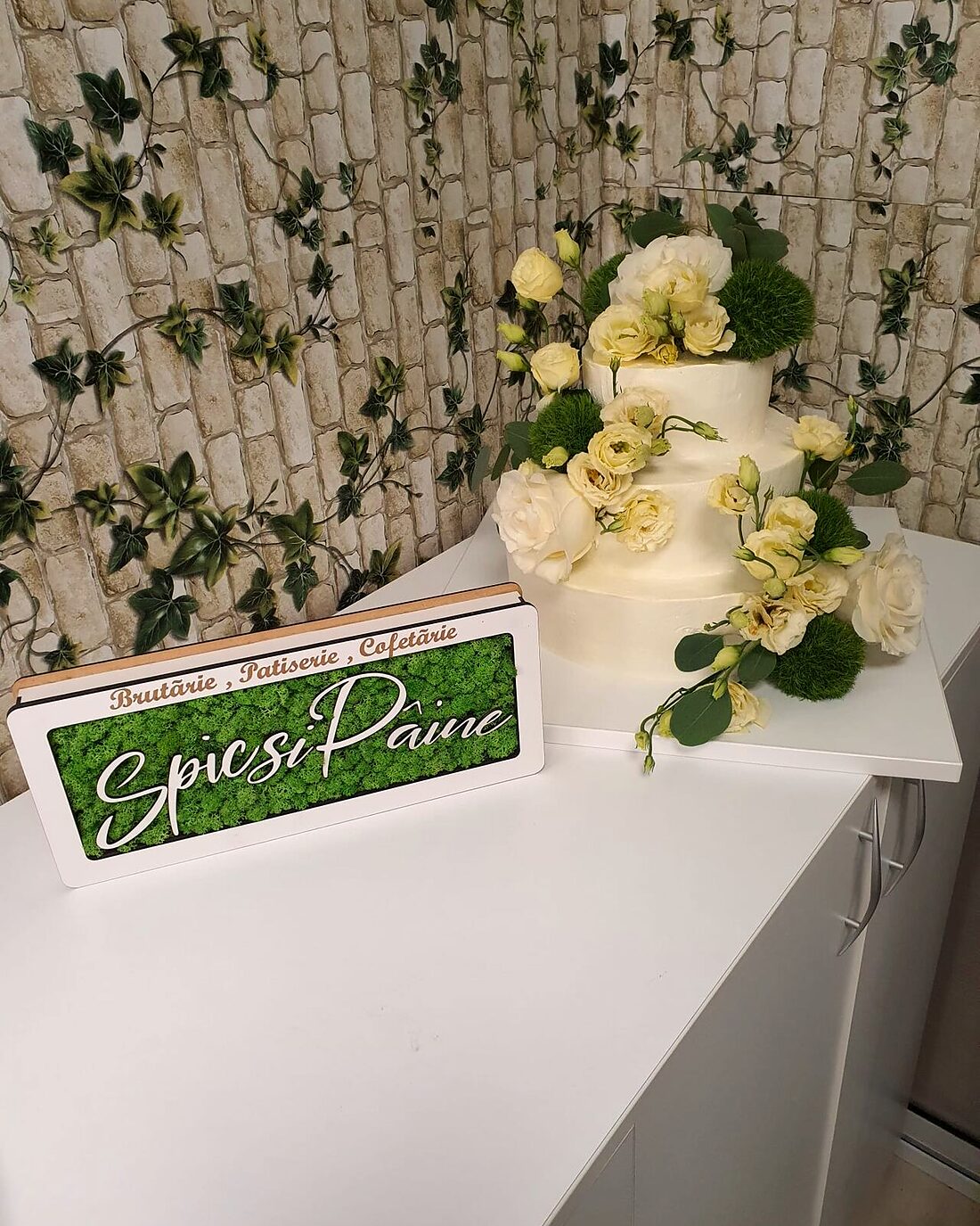 Tort de nuntă alb cu flori de zahăr, în fața panoului verde Spic și Pâine